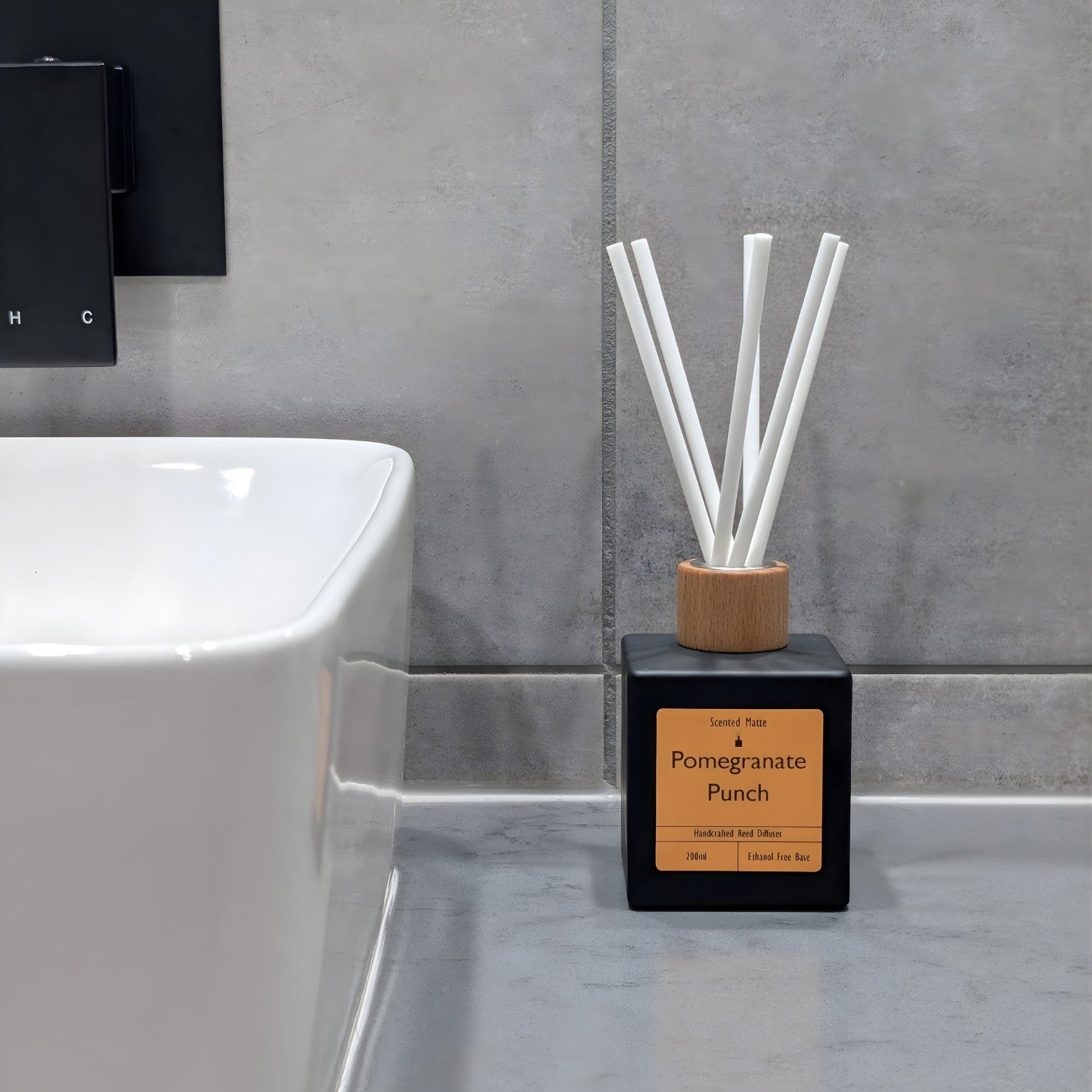 A minimalist matte black reed diffuser with orange label, wooden cap and white fiber reeds, sitting on a grey stone vanity next to a white ceramic sink and grey tiled wall.
