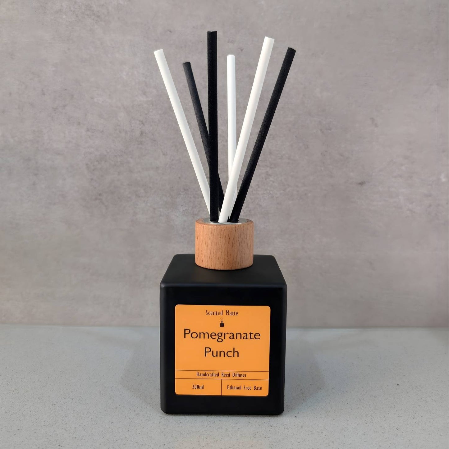 A minimalist matte black square reed diffuser with an orange label, light wood cap and six black/white fiber reeds, set against a grey background.