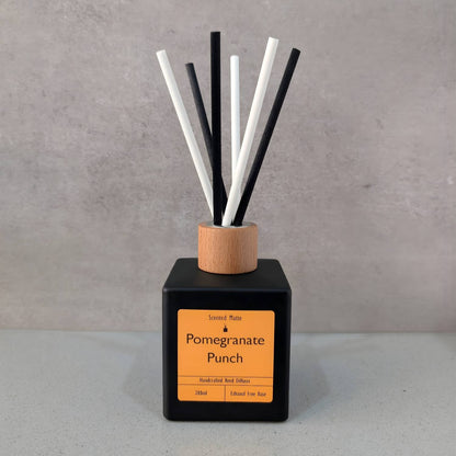 A minimalist matte black square reed diffuser with an orange label, light wood cap and six black/white fiber reeds, set against a grey background.