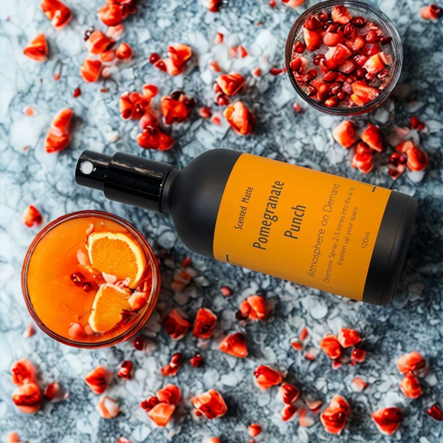 A minimalist matte black room spray bottle with a vibrant orange label, surrounded by ruby red pomegranate seeds and a glass of punch.