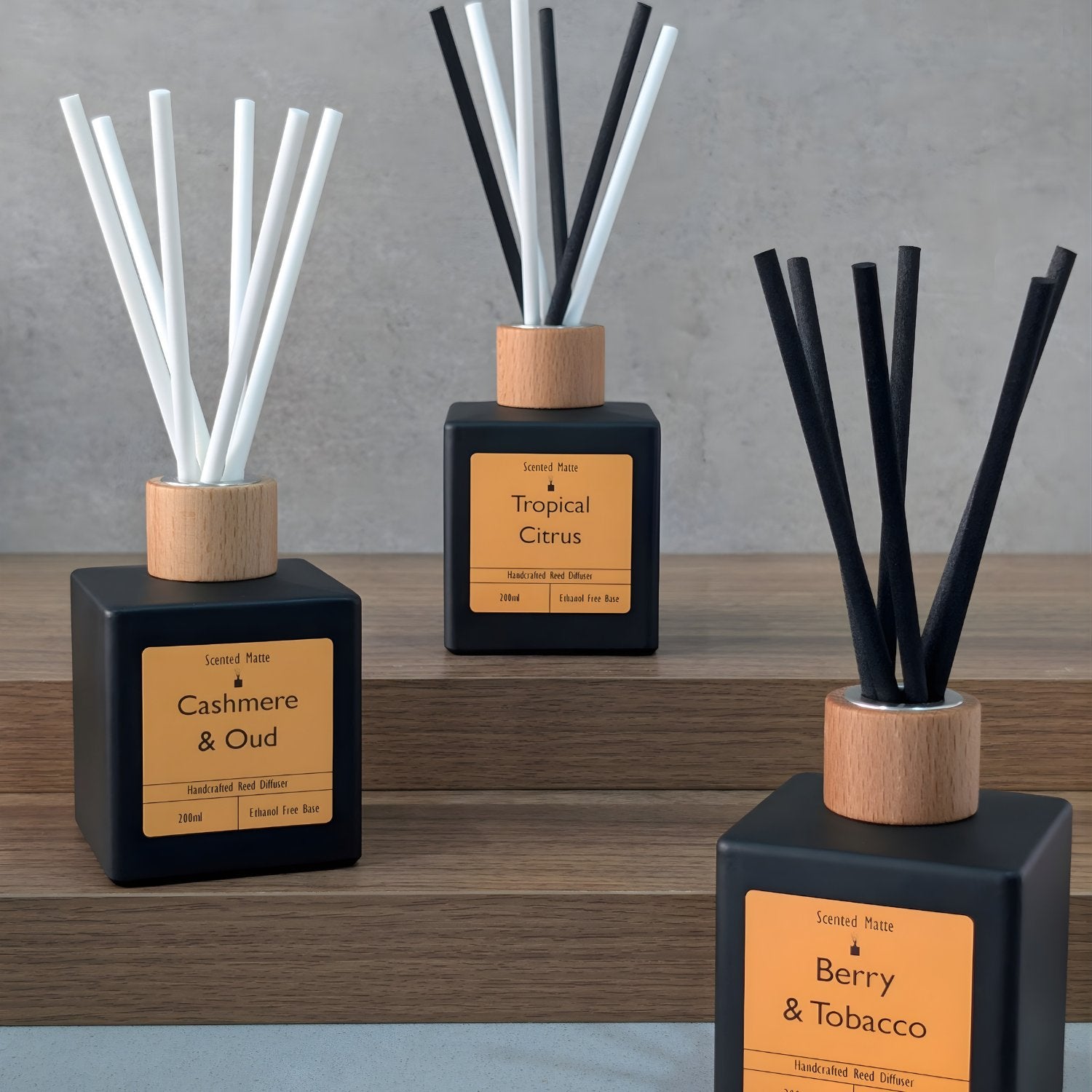 Three Matte black reed diffusers with a wooden cap, showcasing an array of white, black and mixed reed colours on a tiered wooden surface with a tiled grey background. 