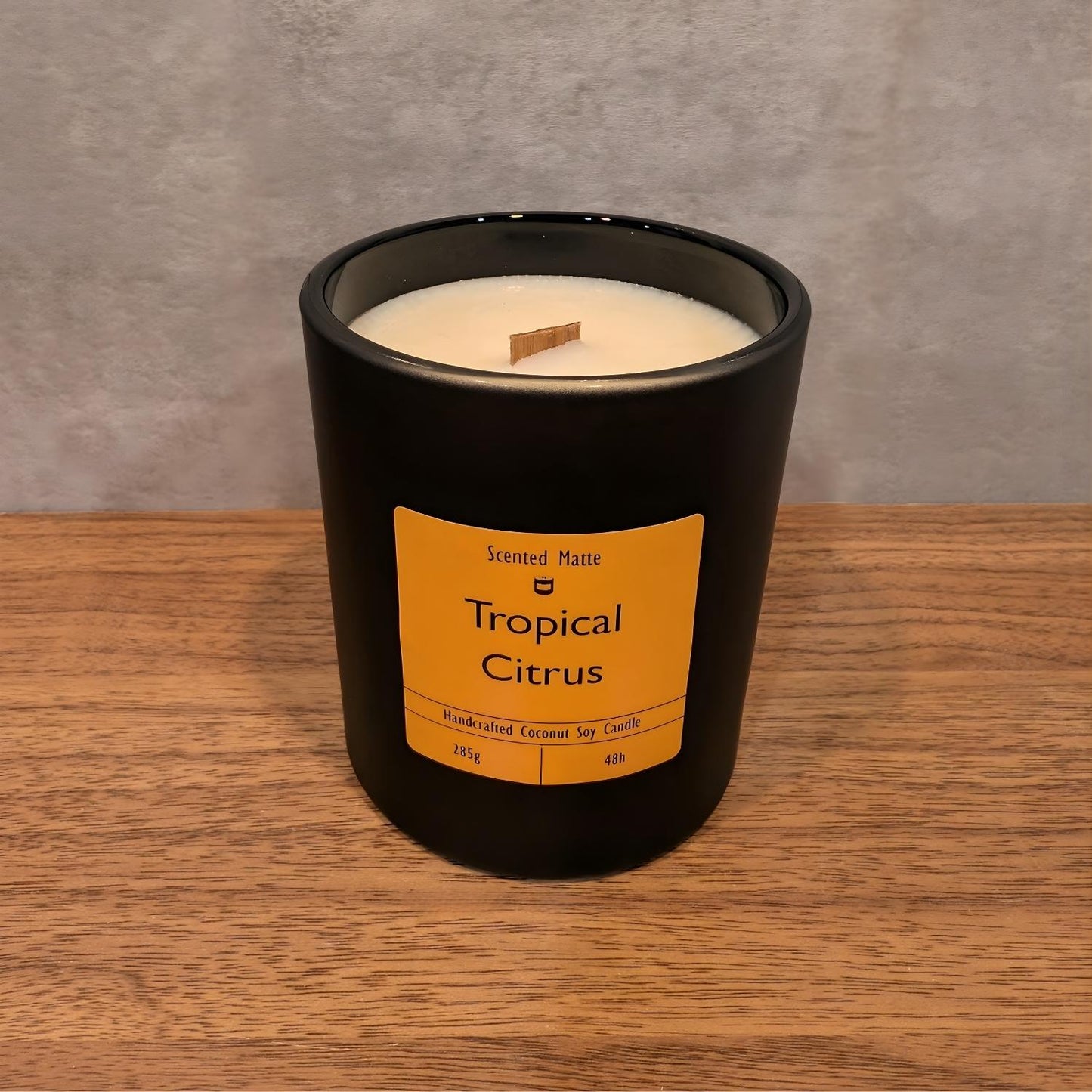 A minimalist matte black candle jar with a wooden wick, sitting on a light oak wood surface against a textured grey background.