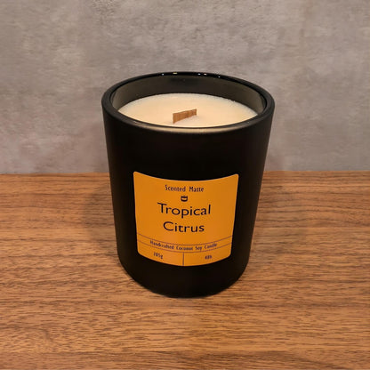 A minimalist matte black candle jar with a wooden wick, sitting on a light oak wood surface against a textured grey background.