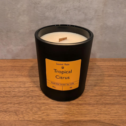 A minimalist matte black candle jar with a wooden wick, sitting on a light oak wood surface against a textured grey background.