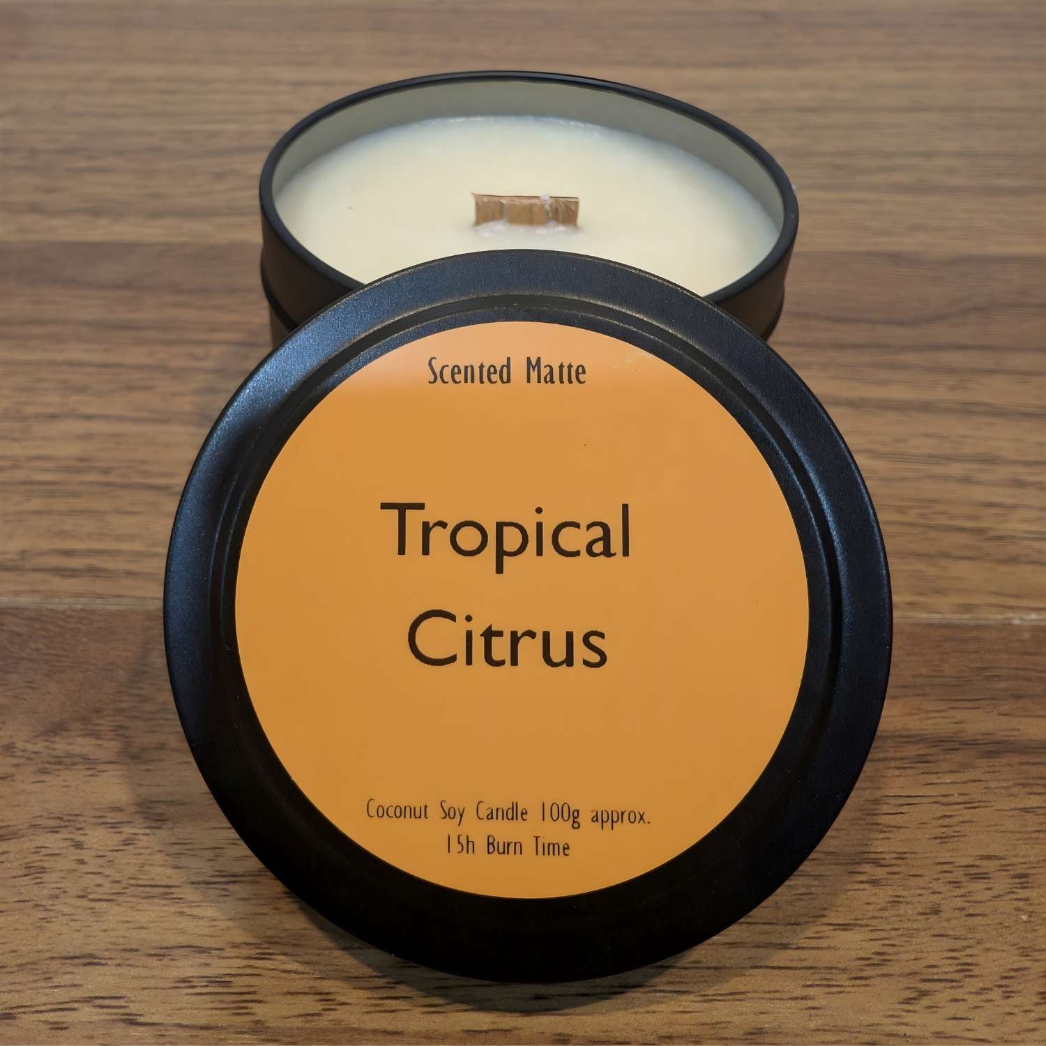 A wooden wick candle in a black tin with orange label on the lid on a wooden surface.