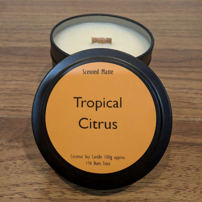 A wooden wick candle in a black tin with orange label on the lid on a wooden surface.