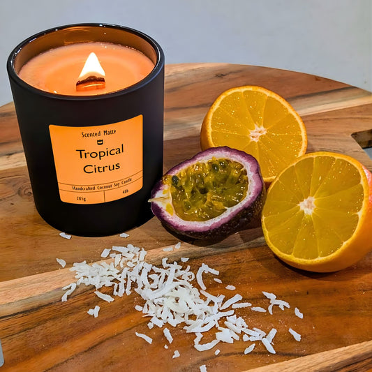 A minimalist matte black candle jar with a lit wooden wick, styled with fresh tangerine and passionfruit halves and coconut flakes.