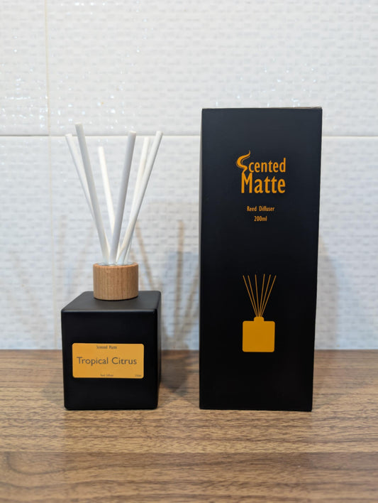Matte black diffuser bottle with white reed sticks and wooden cap, next to its packaging on a wooden surface.