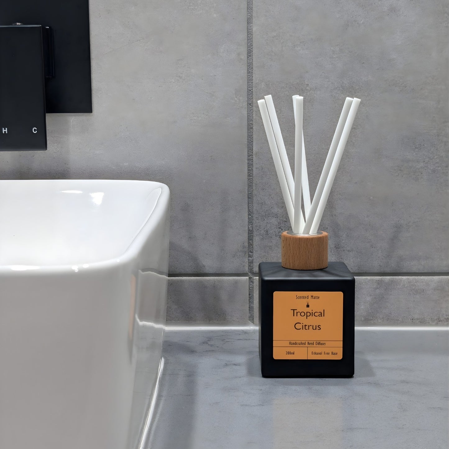 A minimalist matte black reed diffuser with orange label, wooden cap and white fiber reeds, sitting on a grey stone vanity next to a white ceramic sink and grey tiled wall.