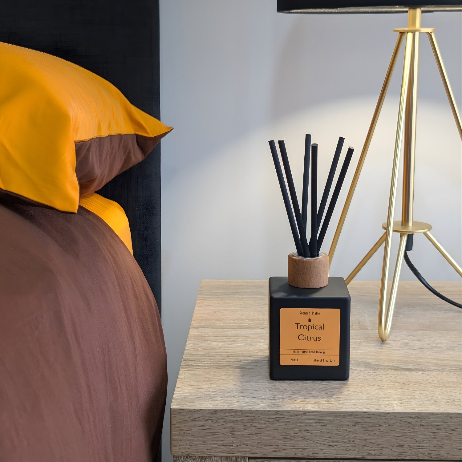 A minimalist matte black reed diffuser with orange label, wood cap and black reeds on a light wood bedside table, next to a gold tripod lamp and a bed with brown and mustard yellow linens.