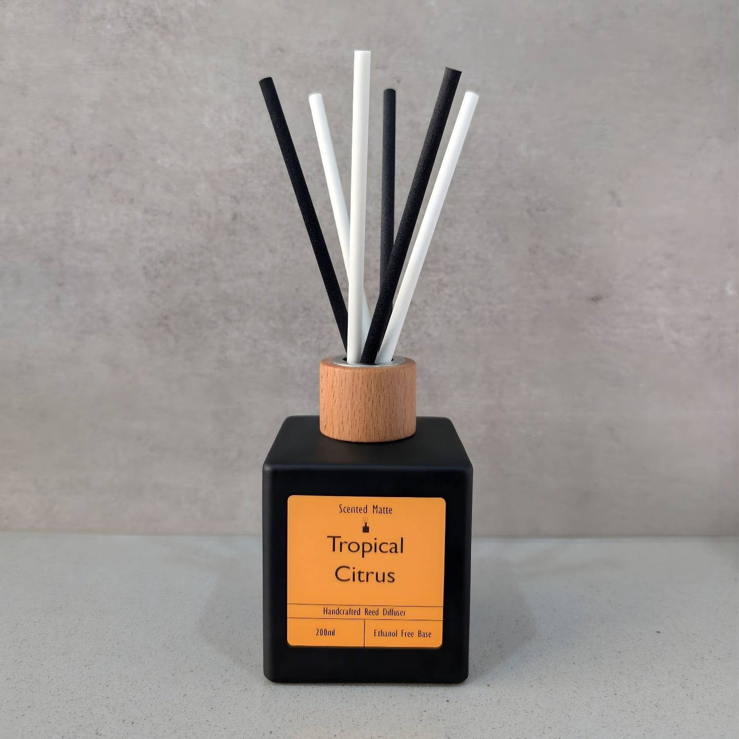 A minimalist matte black square reed diffuser with an orange label, light wood cap and six black/white fiber reeds, set against a grey background.
