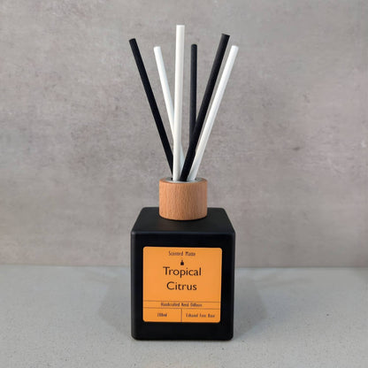A minimalist matte black square reed diffuser with an orange label, light wood cap and six black/white fiber reeds, set against a grey background.