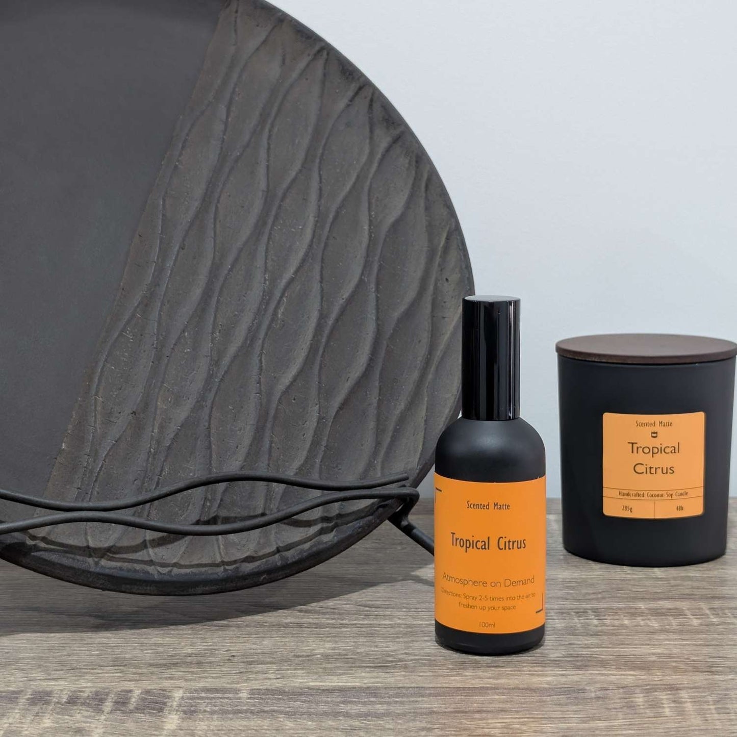 A matte black room spray bottle and a minimalist matte black candle jar, styled next to a large textured charcoal grey decorative plate.
