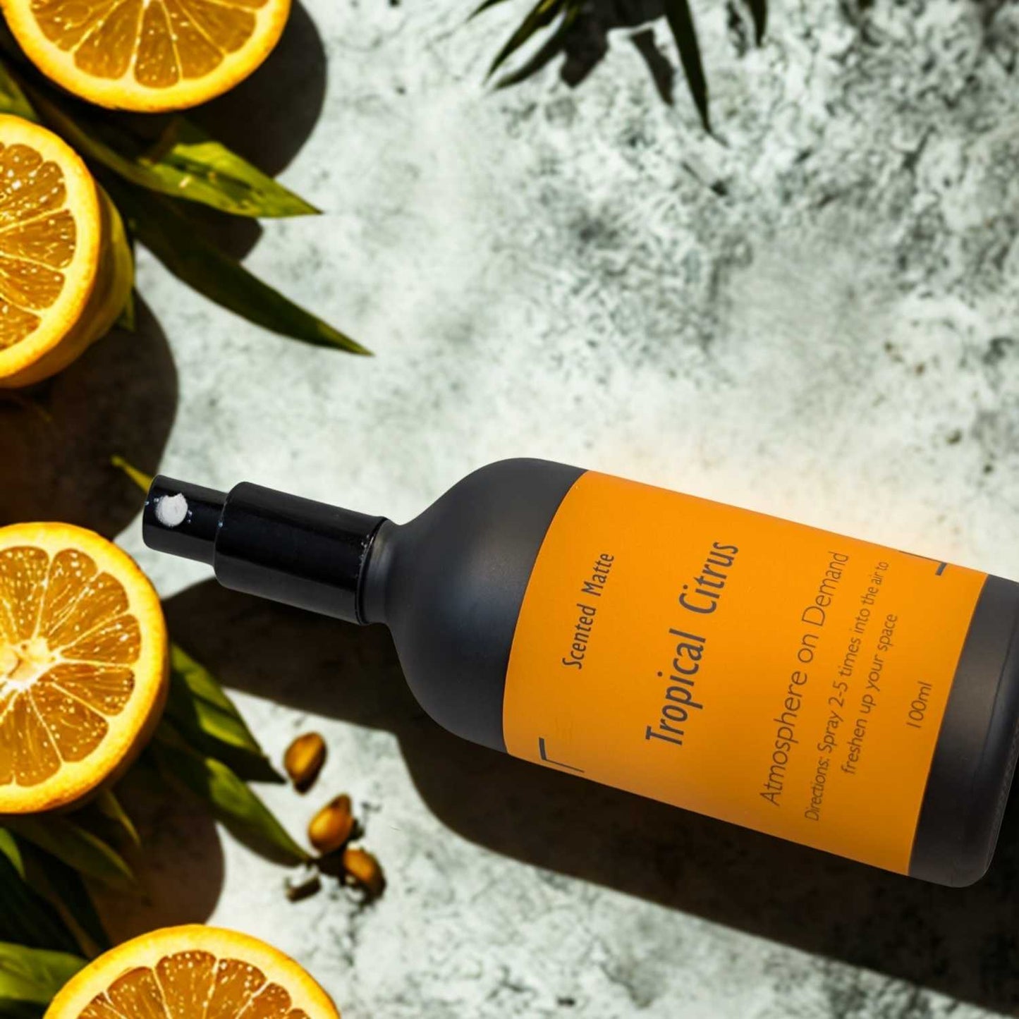 A minimalist matte black room spray bottle with a vibrant orange label, styled with fresh lime halves and vibrant green tropical palm leaves.