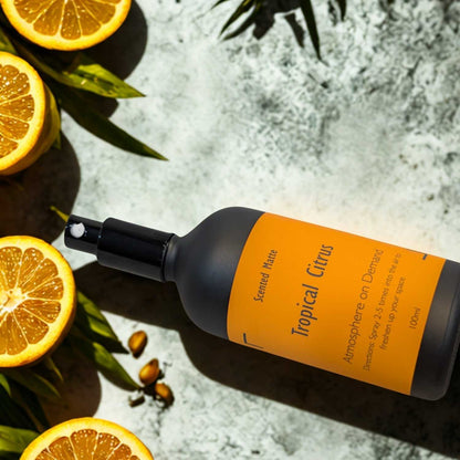 A minimalist matte black room spray bottle with a vibrant orange label, styled with fresh lime halves and vibrant green tropical palm leaves.