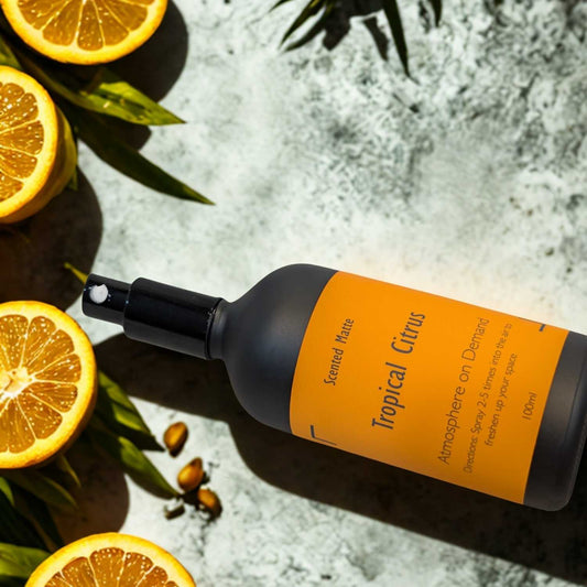A minimalist matte black room spray bottle with a vibrant orange label, styled with fresh lime halves and vibrant green tropical palm leaves.