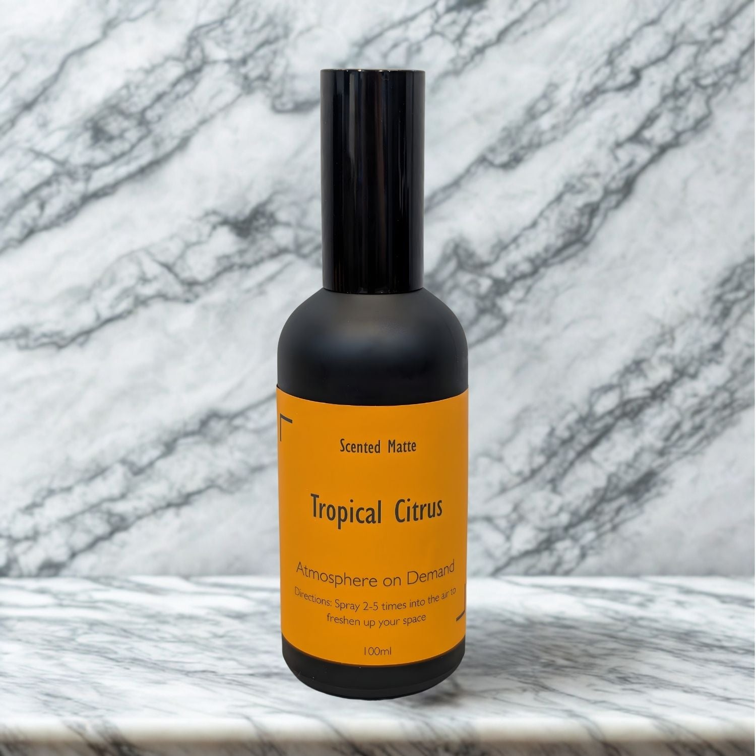 A minimalist matte black 100ml room spray bottle with a vibrant orange label, centered on a white marble surface.