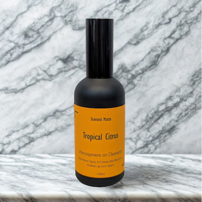 A minimalist matte black 100ml room spray bottle with a vibrant orange label, centered on a white marble surface.